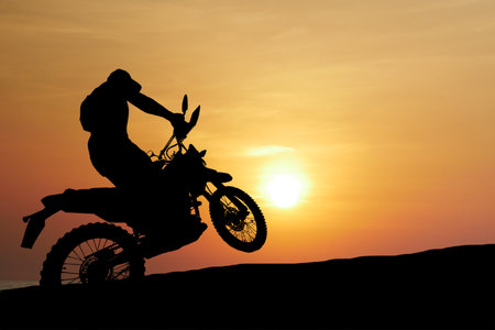 Silhouette of a motorcyclist doing a wheelie on a dirt bike with a vibrant sunset in the background, showcasing adventure and thrill.の写真素材