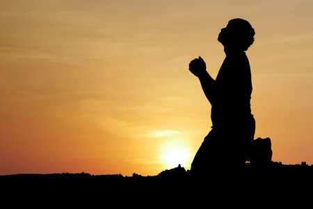 A silhouette of a person kneeling and praying at sunset, with a stunning orange sky and sun setting in the background.の写真素材