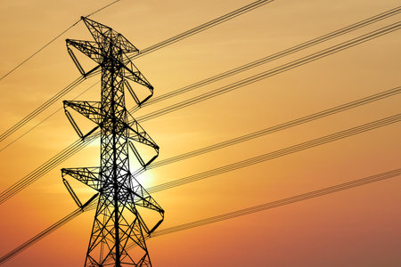 Silhouette of a high voltage transmission tower with power lines against a stunning orange sunset sky.の写真素材