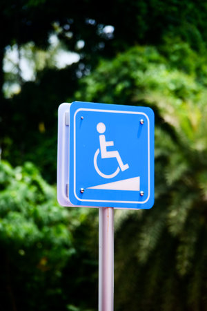 Close-up of a blue wheelchair accessibility sign with a ramp symbol, set against a backdrop of lush green foliage.の写真素材