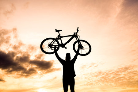 A powerful silhouette of a person triumphantly raising a bicycle against a stunning sunset backdrop with vibrant colors and dramatic clouds.の写真素材