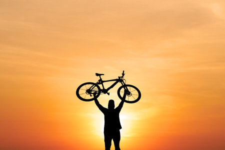 A cyclist raises their bike overhead, silhouetted against a stunning orange sunset, symbolizing achievement and freedom.の写真素材
