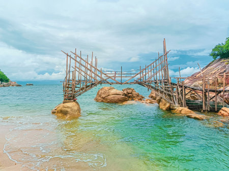 A scenic view of a rustic wooden bridge over turquoise waters on a tropical beach, surrounded by rocks and a cloudy sky.の写真素材