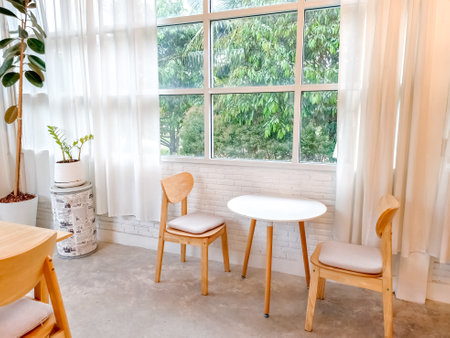 Bright minimalist cafe interior featuring wooden chairs, a small round table, and large windows with lush greenery outside.の写真素材