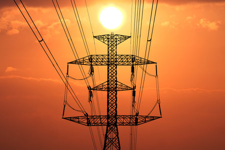 High voltage power line tower silhouetted against a vivid orange sunset, capturing the essence of energy and technology.の写真素材