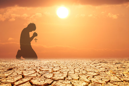 Silhouette of a person kneeling with a cross on cracked earth at sunset, symbolizing faith and hope in a barren landscape.の写真素材