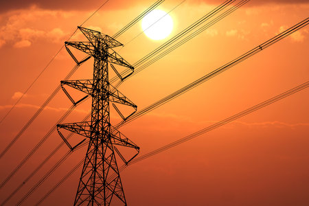 Silhouette of a power line tower against a vivid orange sunset, capturing the interplay of technology and nature.の写真素材