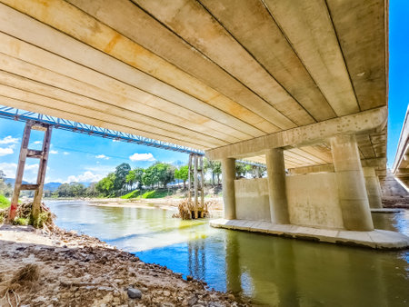 View from beneath a concrete bridge over a calm river, showcasing clear skies and vibrant greenery.の写真素材