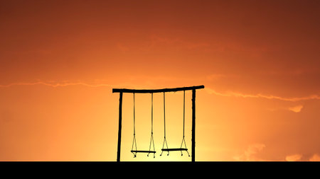 A peaceful silhouette of a swing set with a stunning orange sunset backdrop, evoking nostalgia and tranquility.の写真素材