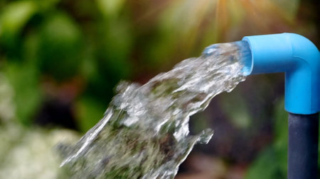 Clear water streaming from a blue PVC pipe in a lush garden setting, illuminated by sunlight.の写真素材
