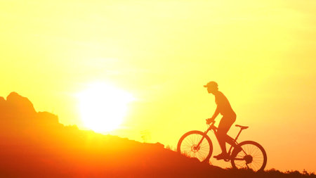 A cyclist silhouetted against a vivid sunset, riding uphill with a glowing sun in the background, capturing the essence of adventure and freedom.の写真素材
