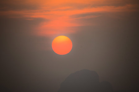 Captivating sunset featuring a vivid orange sun setting behind a silhouetted landscape with a dramatic sky.の写真素材