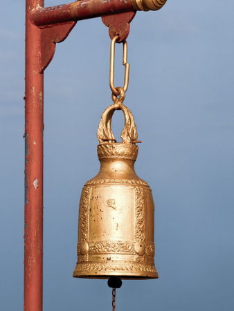 A beautifully crafted golden bell with intricate designs, suspended from a rustic metal frame against a clear blue sky.の写真素材