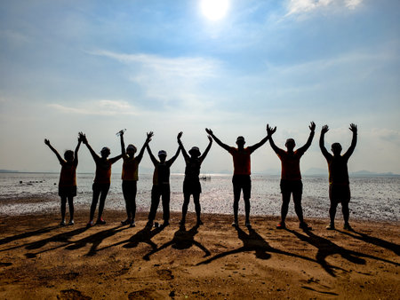 A group of people silhouetted against the sun, celebrating with raised arms on a sandy beach.の写真素材