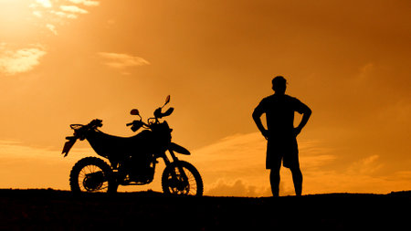 A striking silhouette of a man standing beside a motorcycle, set against a vivid orange sunset sky on a hilltop.の写真素材