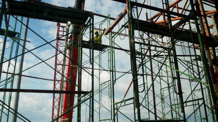 Construction worker on scaffolding at a building site, surrounded by metal framework under a blue sky.の写真素材