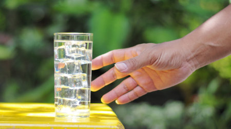 A hand reaches for a glass of ice water on a sunny day, set against a lush green background, symbolizing refreshment and hydration.の写真素材