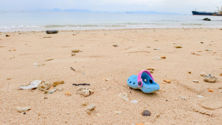 A single blue sandal rests on a sandy beach, surrounded by shells and pebbles, with the ocean and a...
