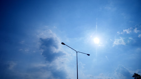 Street lamp silhouette against a vibrant blue sky with sunlight and scattered clouds.の写真素材