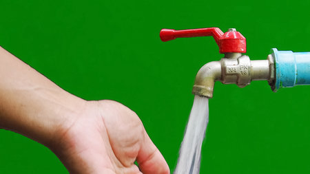 Close-up of a hand reaching for water flowing from a red-handled outdoor faucet, set against a bright green backdrop.の写真素材
