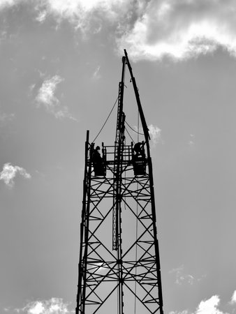 Black and white image of construction workers silhouetted on a tall tower against a cloudy sky.の写真素材