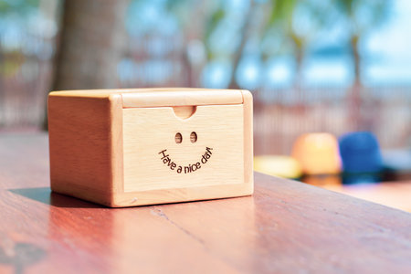 Wooden box with smiley face and "Have a Nice Day" message on a table, set against a sunny, blurred outdoor background.の写真素材
