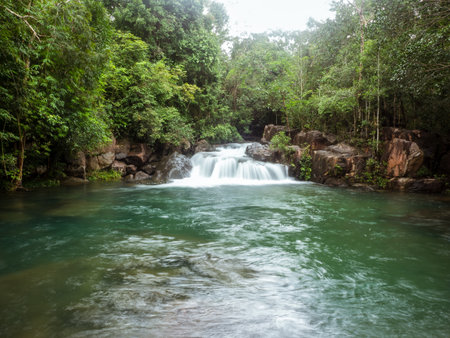A serene tropical waterfall cascading over smooth rocks into a crystal-clear emerald pool surrounded by lush green forest. The gentle stream reflects sunlight filtering through theの写真素材