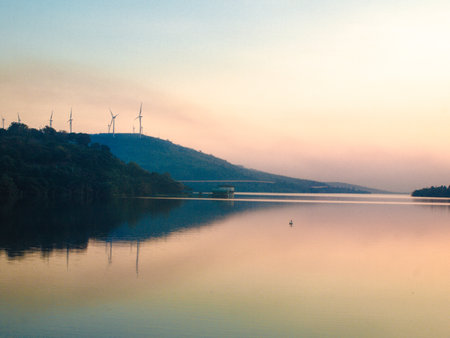 A calm reservoir reflecting the pastel tones of the sky at dawn with wind turbines on the hillside in the background.の写真素材