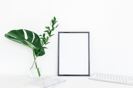 Desk with photo frame, notebook, computer, green leaves. Creative workspace. Front view, copy space, mock upの写真素材