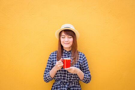smiling woman wear in dress and hat holding red coffee cup on yellow cement wall background, thinking, imaginationの写真素材