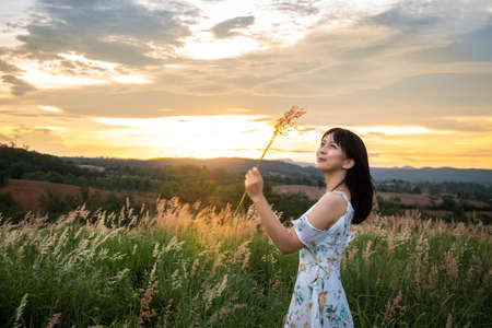 The girl wearing a white dress, stand in the middle of the grass and Hand holding grass with beautiful white flowers with a relaxed and happy mood on mountain in sun set time.の写真素材