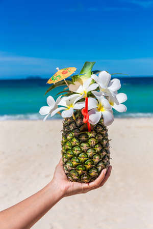 Tropical exotic pineapple cocktail with plumeria on hand at the beach with white sandy and blue seaの写真素材