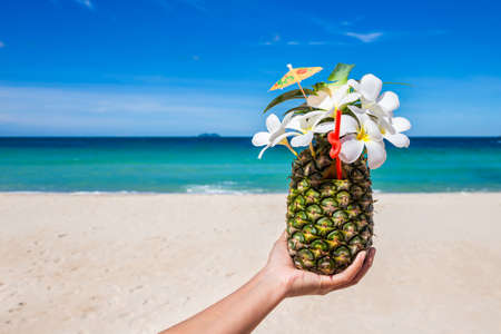 Tropical exotic pineapple cocktail with plumeria on hand at the beach with white sandy and blue seaの写真素材