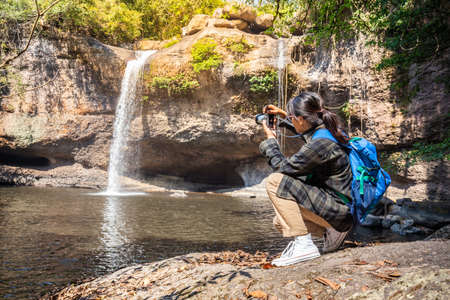 Freedom traveler woman sitting waterfall front on stone with bag and Taking a picture by camera enjoying a beautiful nature. Travel conceptの写真素材