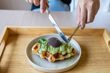 Hands holding hands and a fork prepare to cut green tea waffles in a white plate on a traditional...