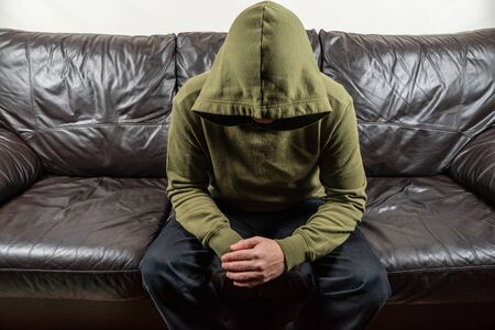 Man in dark green hoodie with unrecognizable face sitting on broun leather sofaの写真素材