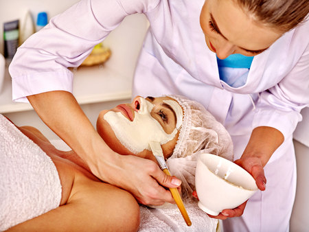 Woman middle-aged take facial and neck clay mask in spa salon. Beautician hold cup and brush.の写真素材