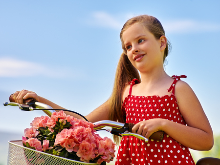 Bikes cycling girl. Child girl wearing white polka dots red dress rides bicycle with pink flowers basket.の写真素材