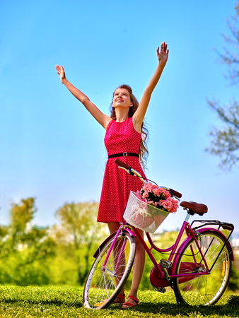 Bikes bicycle girl. Teenager girl wearing red polka dots dress looking up and hand up keeps bicycle with flowers basket. Lot of green tree and blu sky in park.の写真素材
