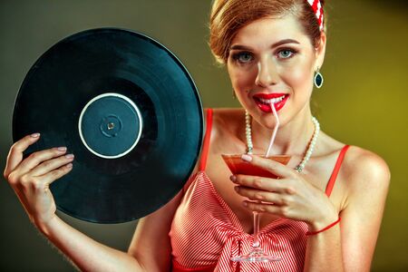 Girl in pin-up style keeps vinyl record and drink martini cocktail . Pin-up retro female style. Girl...
