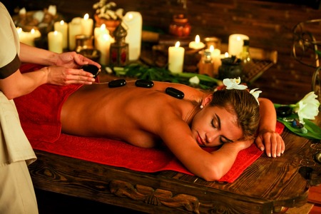 Young woman lying on wooden spa bed. Lastone therapy massage in spa salon. On girl back are stones. Girl on candles background in massage spa salon.の写真素材
