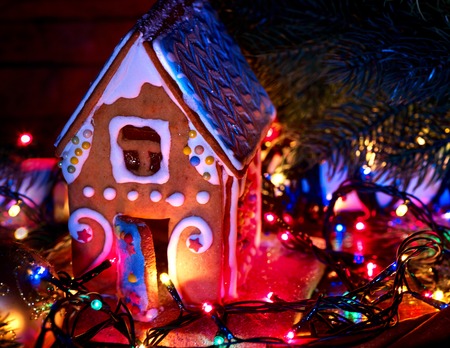 Gingerbread houses with christmas lights string on wooden table. Close up Xmas cookies decorating frosting a cake indoor. Light effect for festive dinner. Nobody on pictute.の写真素材