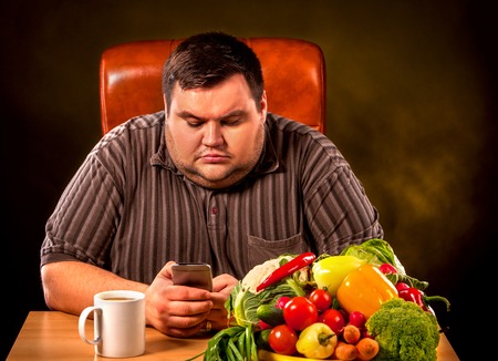 Diet fat man eating healthy food . Healthy breakfast with vegetables for overweight person. Male trying to lose weight but orders fast pizza by phone. Concept on black background.の写真素材