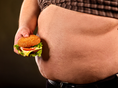 Man belly fat with hamberger fast food . Breakfast for overweight person. Cropped shot of junk meal leads to obesity. Person reduced consumption of hamburgers on black background.の写真素材