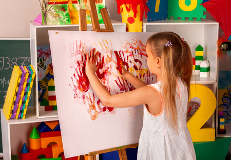 Child painting finger on easel. Girl learn paint in class school. Kindergarten interior on background. Picture with prints of childrens hands. Free childrens creativity.の写真素材