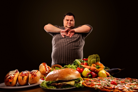 Diet fat man who makes choice between healthy and unhealthy food . Overweight male with hamburgers, french fries and vegetables trays trying to lose weight first time .Abundance of food concept.の写真素材