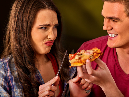 Couple eating in fast food restaurant. Man and woman eat slice pizza with knife and fork. Friends holding junk on dark background. Girl against the use of semi-finished products idea.の写真素材