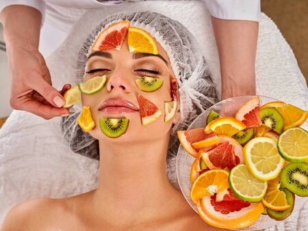 Facial mask from fresh fruits for woman. Beautician apply slices of avocado, grapefruit and kiwi Girl in medical hat lying on spa bed for skin care procedure in salon. Female enjoys procedure.の写真素材