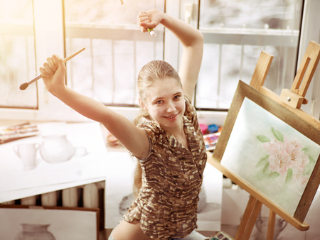 Artist painting on easel in studio. Happy authentic girl paints with brush in morning sunlight dawn light toning. School Indoor interior . Top view of drawing of spring flowers on window background.の写真素材
