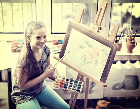 Artist painting on easel and palette in studio. Authentic girl paints with oil brush morning sunlight dawn light toning window background. Drawing of spring flowers. Stylization of photo under film.の写真素材
